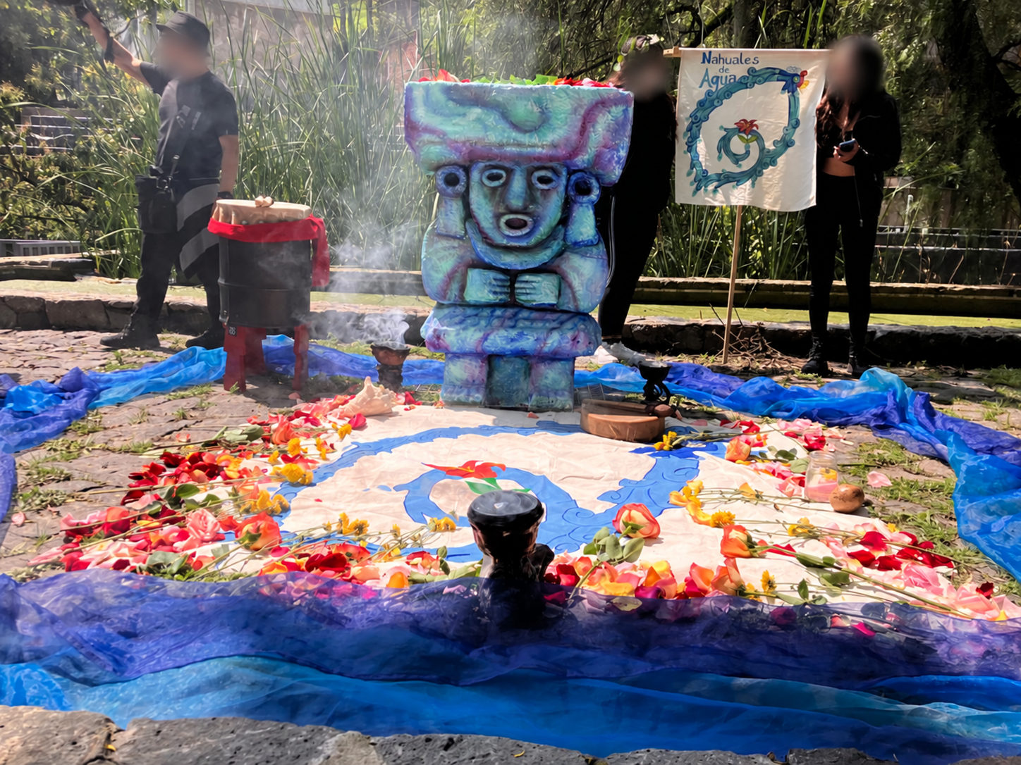 Outdoor gathering with a sculpture, flowers, blue fabrics and a Nahuales de Agua banner. All three faces are blurred.